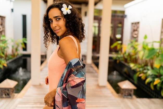 Retrato de verano al aire libre de una linda mujer morena con flores tropicales en los pelos posando en un hotel elegante