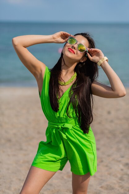 Retrato de verano al aire libre de moda de mujer hermosa morena bastante joven en gafas de sol frescas posando en la soleada playa tropical.