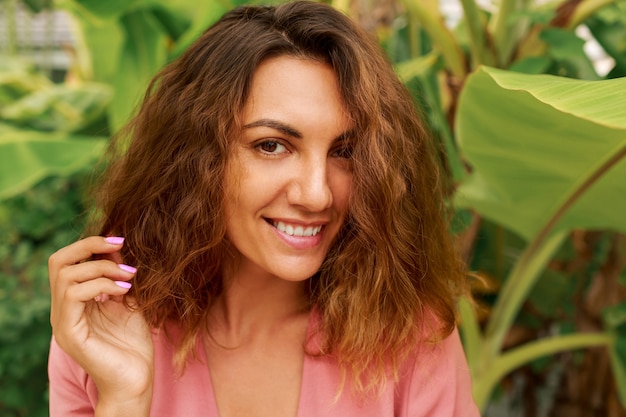 Retrato de verano al aire libre de seductora mujer morena con pelos ondulados en vestido rosa posando en tropical