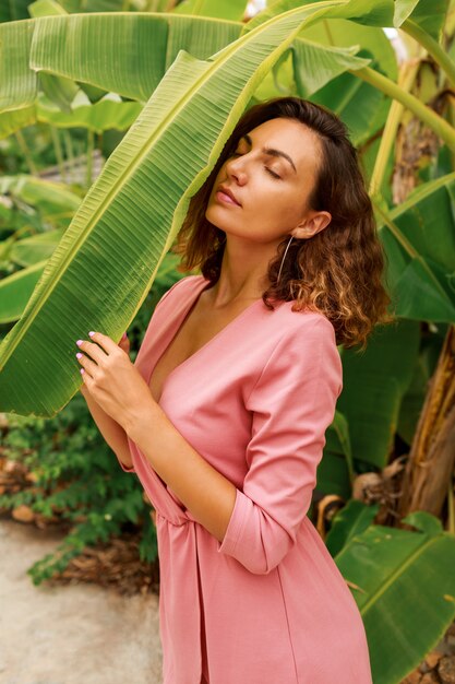 Retrato de verano al aire libre de seductora mujer morena con pelos ondulados en vestido rosa posando