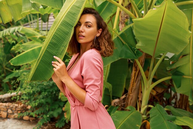 Retrato de verano al aire libre de seductora mujer morena con pelos ondulados en vestido rosa posando