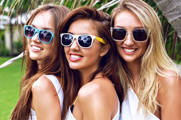 Retrato de verano de árbol joven mujer bonita sexy de cerca, vestida con blusas blancas de moda casual simple y gafas de sol brillantes, sonriendo divirtiéndose de vacaciones en un país tropical.