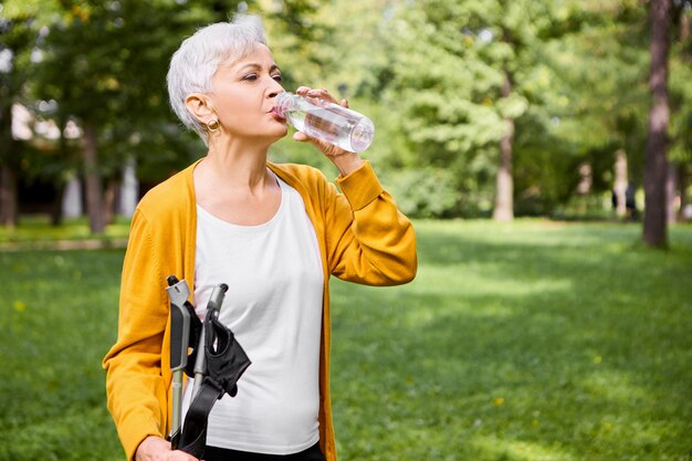 Retrato de verano de cansada mujer caucásica de pelo gris en sus sesenta años bebiendo agua de una botella de plástico, refrescándose después de la actividad física, posando al aire libre con bastones de marcha nórdica