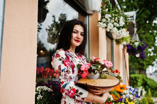 Retrato de verano de una chica morena con gafas rosas y sombrero contra la tienda de flores