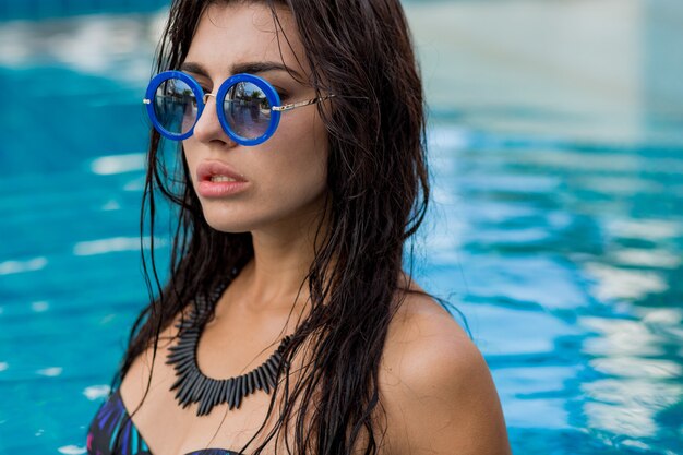 Retrato de verano de hermosa modelo sexy en traje de baño negro y elegante collar posando en la piscina. Vacaciones y ambiente tropical.