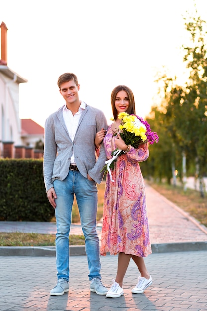 Retrato de verano de una linda pareja increíble caminando por la calle, puesta de sol de campo, ropa elegante, flores, cita romántica, amantes lindos caminando por la calle.