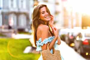Foto gratuita retrato de verano de moda de estilo de vida de elegante joven modelo magnífico posando en la calle, luz del sol brillante de la tarde, elegante vestido azul femenino y bolso de paja, experiencia de viaje.