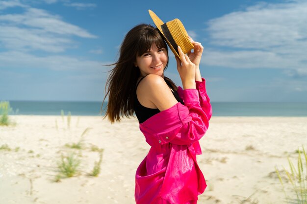 Retrato de verano de moda mujer bonita con sombrero de paja posando en la playa tropical de cerca. Vistiendo traje de vacaciones rosa.