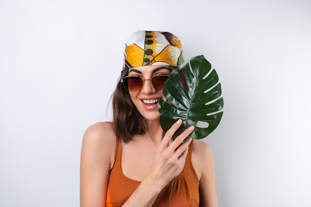 Retrato de verano de una mujer joven en un pañuelo de traje de baño deportivo y gafas de sol con hoja de palma de monstera
