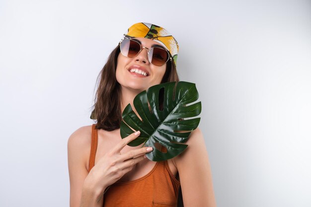 Retrato de verano de una mujer joven en un pañuelo de traje de baño deportivo y gafas de sol con hoja de palma de monstera