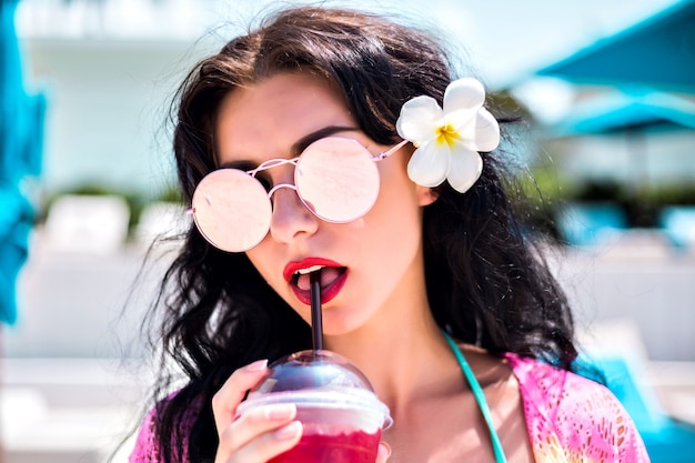 Retrato de verano en tonos soleados brillantes de mujer morena feliz relajante en vacaciones exóticas tropicales, bebiendo limonada, gafas de sol de moda y flor de Tiara natural en sus cabellos.