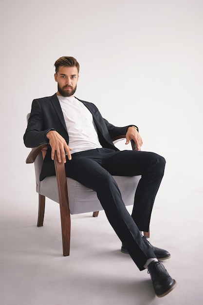 Retrato vertical aislado del exitoso jefe masculino europeo joven con estilo guapo con barba recortada difusa con ropa de hombre de moda relajante en el sillón y mirando con mirada seria