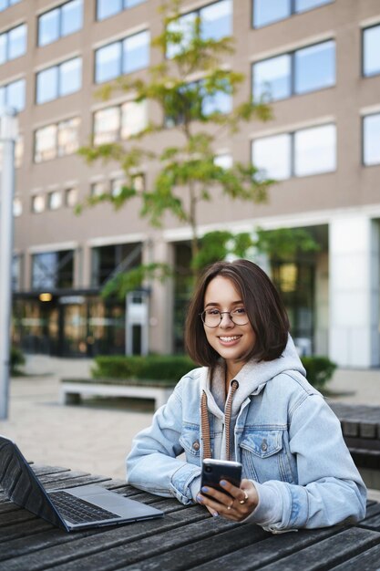 Retrato vertical de una chica caucásica feliz con chaqueta de mezclilla y anteojos usando un teléfono móvil trabajando en un informe fuera de la oficina en un proyecto de escritura de clima perfecto portátil riendo y sonriendo