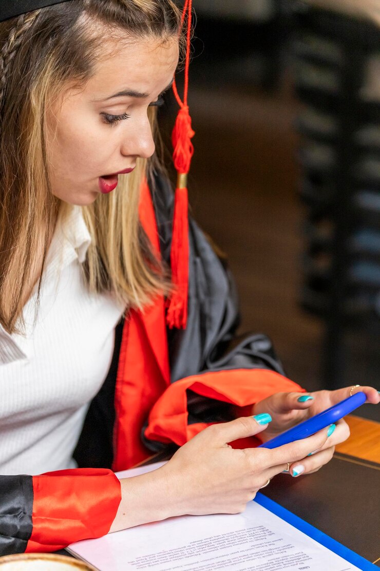 Foto gratuito retrato vertical de una dama sorprendida con capa de graduación mirando su teléfono