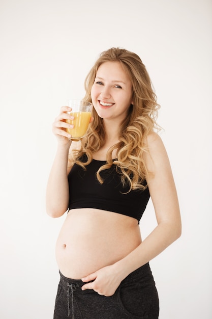 Retrato vertical de feliz alegre mujer embarazada europea con cabello rizado rubio en ropa de casa con el vientre abierto