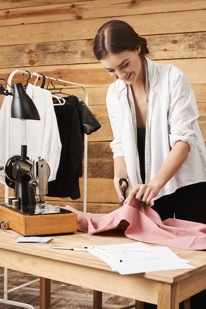 Foto gratuita retrato vertical de feliz entusiasta sastre mujer sonriendo mientras disfruta de su trabajo en el taller, cortando tela con unas tijeras, planeando coser en la máquina de coser nueva paz de la prenda.