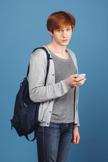 Retrato vertical de guapo joven pelirroja estudiante en traje casual con mochila con smartphone en la mano, con expresión triste e insegura.