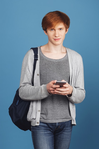 Retrato vertical de hermoso chico serio joven estudiante con cabello rojo en traje casual con mochila negra conversando con su novia por teléfono, con expresión tranquila