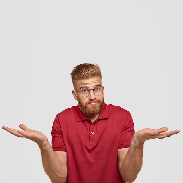 Retrato vertical de hombre astuto dudoso con expresión de desconcierto despistado, siente incertidumbre sobre algo, usa gafas redondas y camiseta roja, se para contra la pared blanca con espacio en blanco