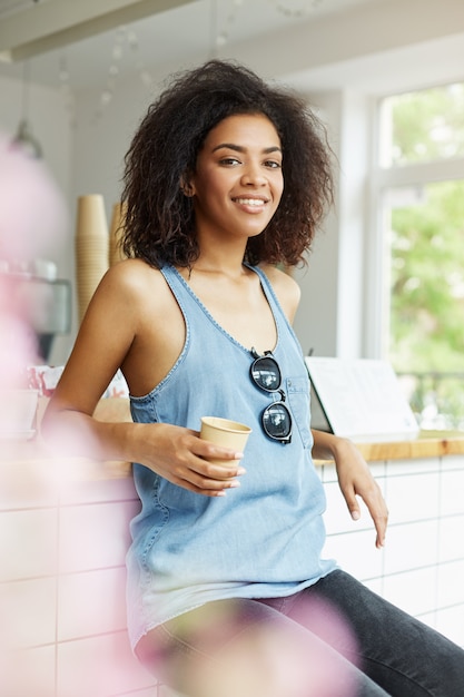 Retrato vertical de joven apuesto alegre alegre chica de piel oscura africal con cabello ondulado en camisa azul sentado en la cafetería, bebiendo café con leche, sonriendo, mirando a la cámara con feliz y rela