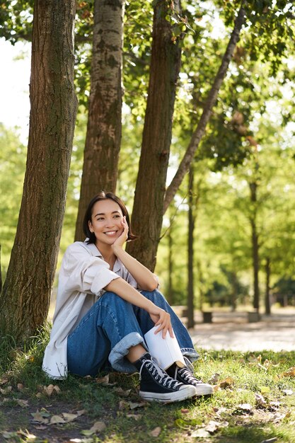 Foto gratuita retrato vertical de una joven asiática leyendo su libro en el parque con un aspecto romántico y sonriente inclinado