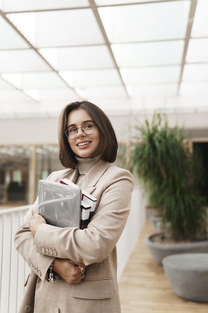 Retrato vertical de una joven empleada o empresaria exitosa en la tutora de la oficina que lleva una computadora portátil y cuadernos mirando hacia otro lado con una sonrisa satisfecha como su trabajo