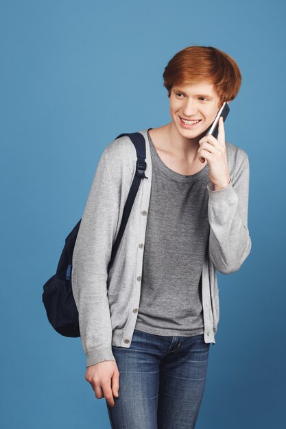 Retrato vertical de joven estudiante guapo con el pelo rojo con traje casual y mochila sonriendo, hablando por teléfono inteligente con un amigo en su camino a la universidad.