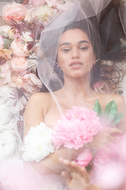 Retrato vertical de una joven modelo acostada bajo tul blanco y sosteniendo un ramo de flores Foto de alta calidad