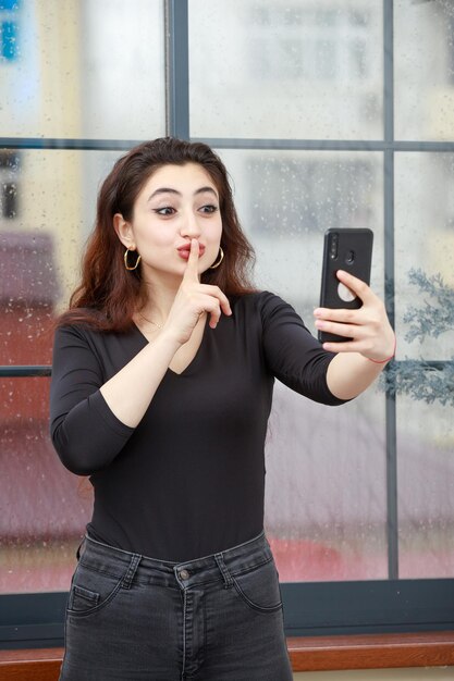 Retrato vertical de una joven sosteniendo el teléfono y haciendo gestos en silencio Foto de alta calidad