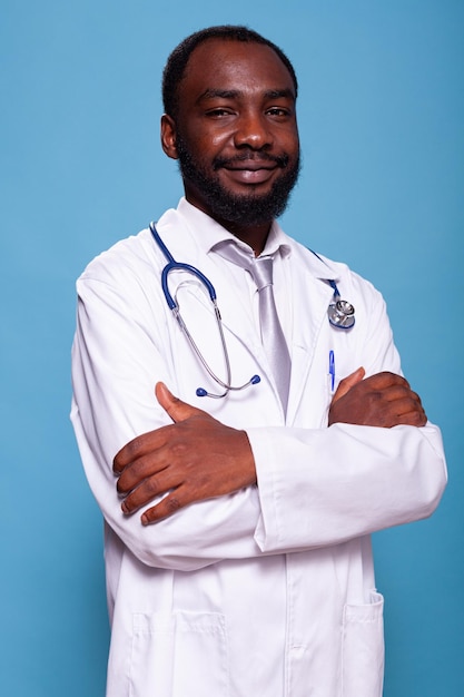 Retrato vertical de un médico afroamericano con estetoscopio posando con confianza usando matorrales. Médico sonriente con uniforme blanco de bata de laboratorio de pie con los brazos cruzados en el estudio.