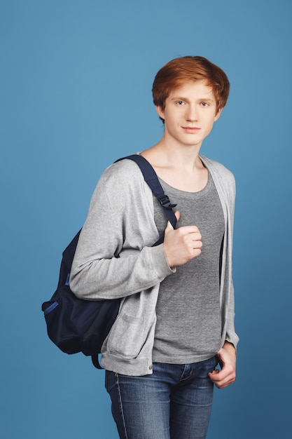 Retrato vertical de serio joven estudiante pelirrojo en traje gris casual con mochila negra, tomados de la mano en el bolsillo, con expresión relajada y segura