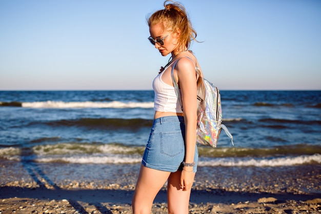Retrato de viaje de estilo de vida al aire libre de joven rubia posando cerca del océano, top corto, mini denim hipster vintage shorty, mochila y gafas de sol, listo para aventuras.