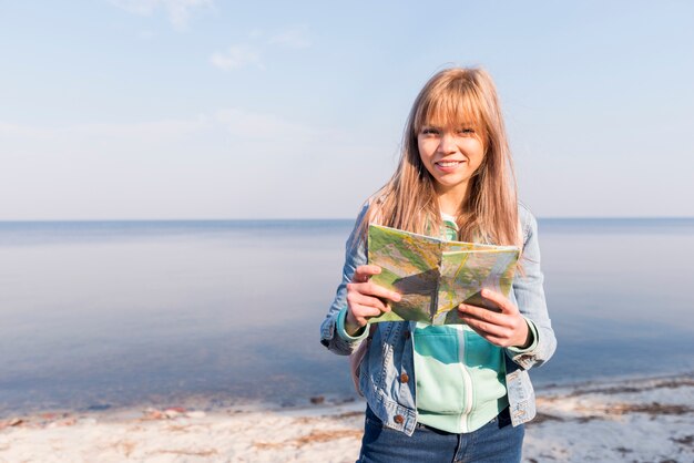 Retrato de un viajero femenino que sostiene el mapa en la mano mirando a la cámara de pie cerca del mar