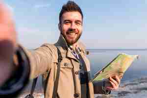 Foto gratuita retrato de un viajero masculino joven sosteniendo un mapa en la mano tomando selfie