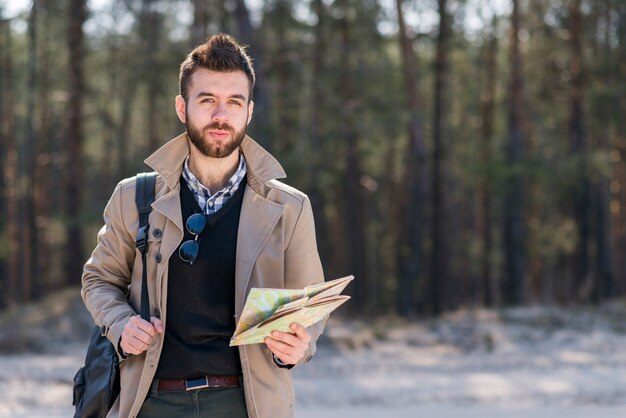 Retrato de un viajero masculino con su mochila en el hombro sosteniendo un mapa en la mano mirando a la cámara