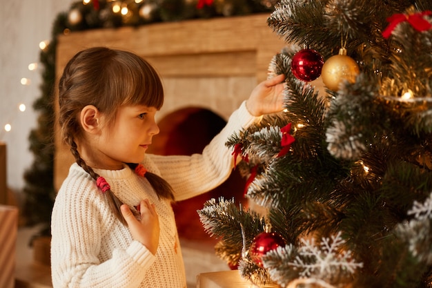 Retrato de vista lateral de la encantadora niña con coletas decorando el árbol de Navidad solo, vistiendo un suéter blanco, de pie en la sala de estar junto a la chimenea.