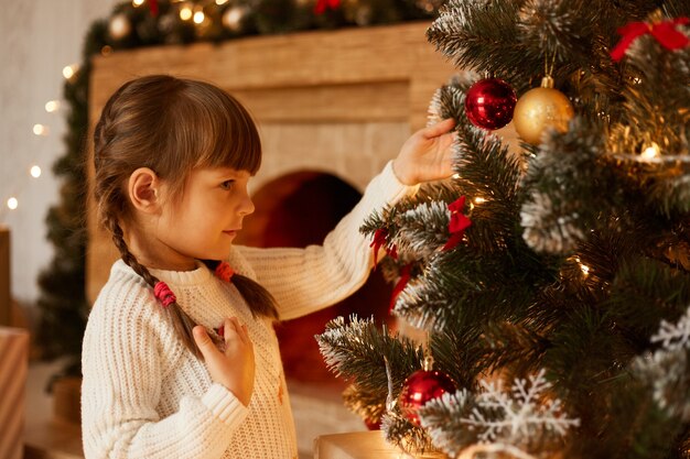 Retrato de vista lateral de la encantadora niña con coletas decorando el árbol de Navidad solo, vistiendo un suéter blanco, de pie en la sala de estar junto a la chimenea.