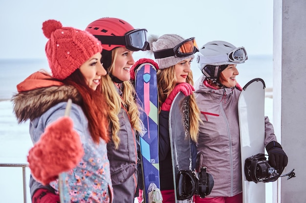 Retrato de vista lateral de una mujer amiga con ropa deportiva de invierno sosteniendo tablas de snowboard y esquís de pie juntas cerca de la costa nevada del mar