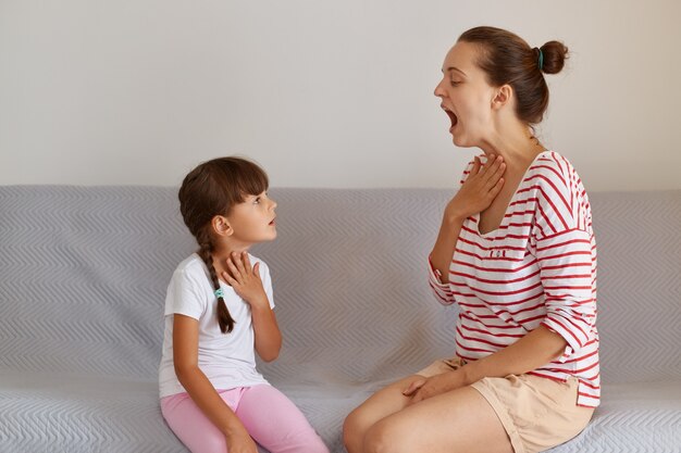 Retrato de vista lateral del patólogo del habla demostrando a un niño pequeño cómo pronunciar correctamente los sonidos, fisioterapeuta profesional que trabaja en los defectos del habla con una niña pequeña en el interior.