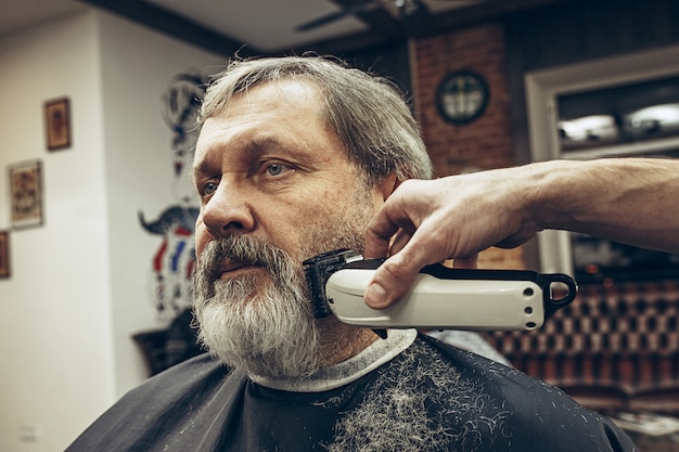 Retrato de la vista lateral del primer del hombre caucásico barbudo mayor hermoso que consigue la preparación de la barba en barbería moderna.