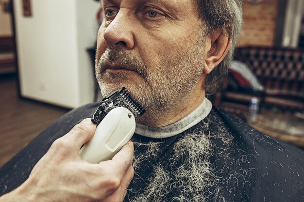 Foto gratuita retrato de la vista lateral del primer del hombre caucásico barbudo mayor hermoso que consigue la preparación de la barba en barbería moderna.