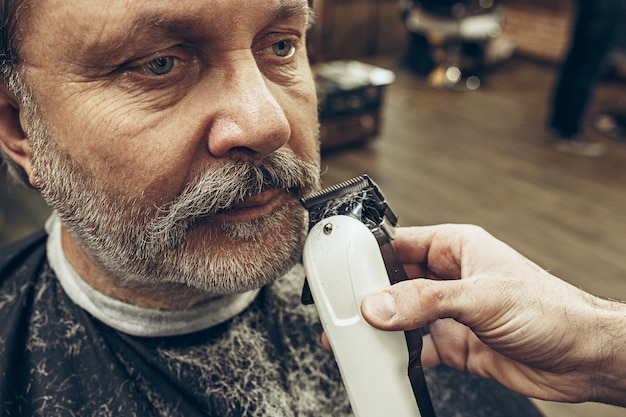 Retrato de la vista lateral del primer del hombre caucásico barbudo mayor hermoso que consigue la preparación de la barba en barbería moderna.