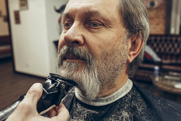 Retrato de la vista lateral del primer del hombre caucásico barbudo mayor hermoso que consigue la preparación de la barba en peluquería moderna.