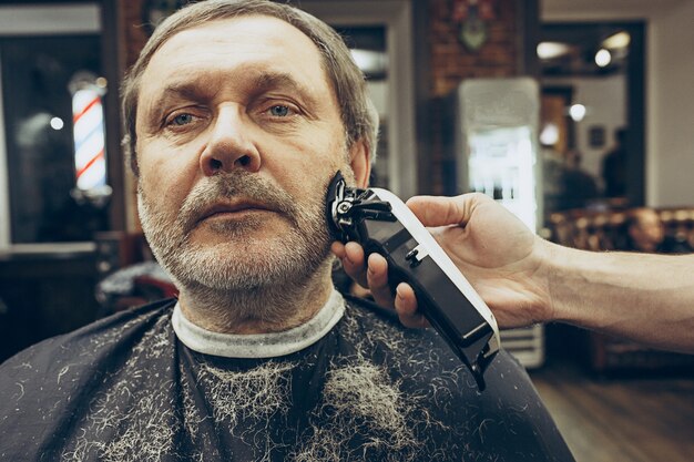 Retrato de la vista lateral del primer del hombre caucásico barbudo mayor hermoso que consigue la preparación de la barba en peluquería moderna.