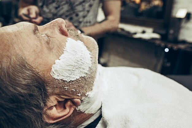 Retrato de la vista lateral del primer del hombre caucásico barbudo mayor hermoso que consigue la preparación de la barba en peluquería moderna.