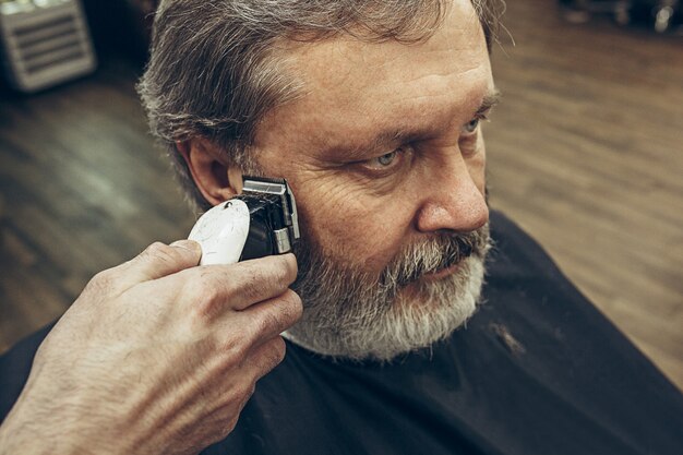 Retrato de vista lateral de primer plano de guapo hombre caucásico barbudo senior conseguir barba aseo en la moderna barbería.