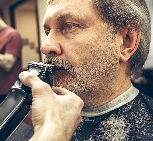 Retrato de vista lateral de primer plano de guapo hombre caucásico barbudo senior conseguir barba aseo en la moderna barbería.