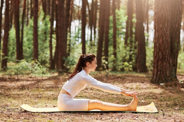 Retrato de vista lateral de ropa deportiva sentado en posición de abdominales al aire libre, entrenamiento de yoga en el bosque, entrenamiento en la naturaleza, relajación y meditación, estilo de vida saludable.