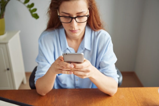 Foto gratuita retrato de vista recortada de linda chica pelirroja enviando mensajes de texto por teléfono celular en el escritorio de madera