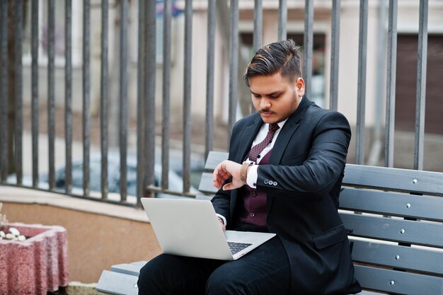 Una reunión importante Elegante hombre de negocios indio con ropa formal sentado en un banco con una computadora portátil y mirando relojes